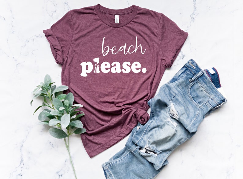 May include: A maroon t-shirt with the words "beach please" in white script and block letters. The shirt is paired with distressed blue jeans and a sprig of green leaves, all arranged on a white marble surface.