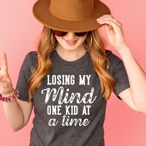May include: A woman wearing a gray t-shirt with the text "Losing my mind one kid at a time" in white. She is wearing a brown hat and sunglasses and has long blonde hair.