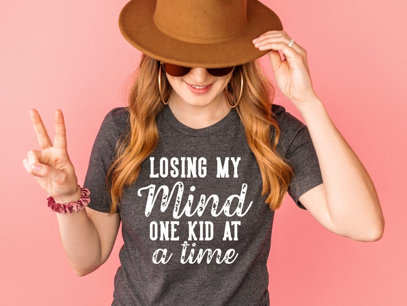 May include: A woman wearing a gray t-shirt with the text "Losing my mind one kid at a time" in white. She is wearing a brown hat and sunglasses and has long blonde hair.