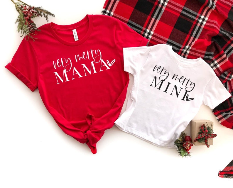 May include: Two red and white Christmas shirts with the text "very merry mama" and "very merry mini" on them. The shirts are laid out on a white surface with a red and black plaid blanket in the background.
