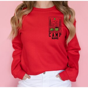 May include: Red long-sleeved sweatshirt featuring a graphic of a camping scene with a tent, mountains, and a mug of coffee. The text "CAMP NAME" is below the graphic.