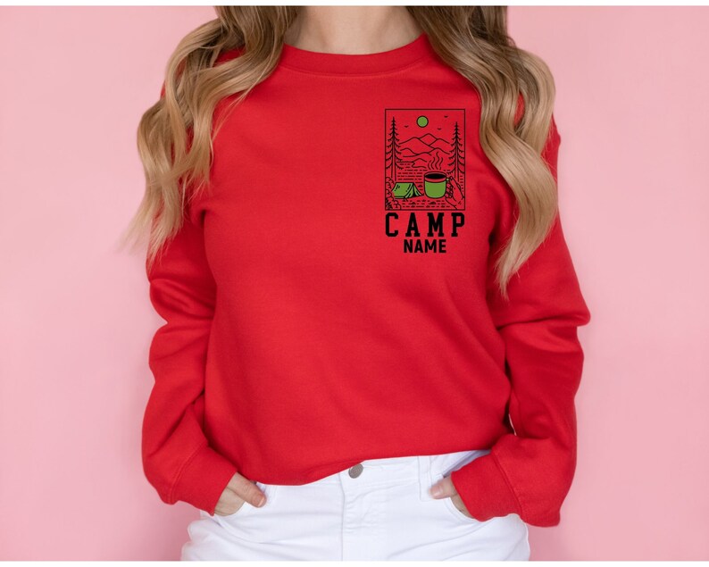 May include: Red long-sleeved sweatshirt featuring a graphic of a camping scene with a tent, mountains, and a mug of coffee. The text "CAMP NAME" is below the graphic.