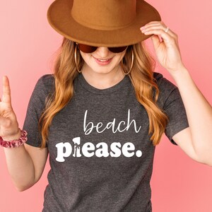 May include: A dark grey t-shirt with the words "beach please" in white script. The person in the image is wearing a brown hat, sunglasses, and gold hoop earrings. A scrunchie is on the wrist.