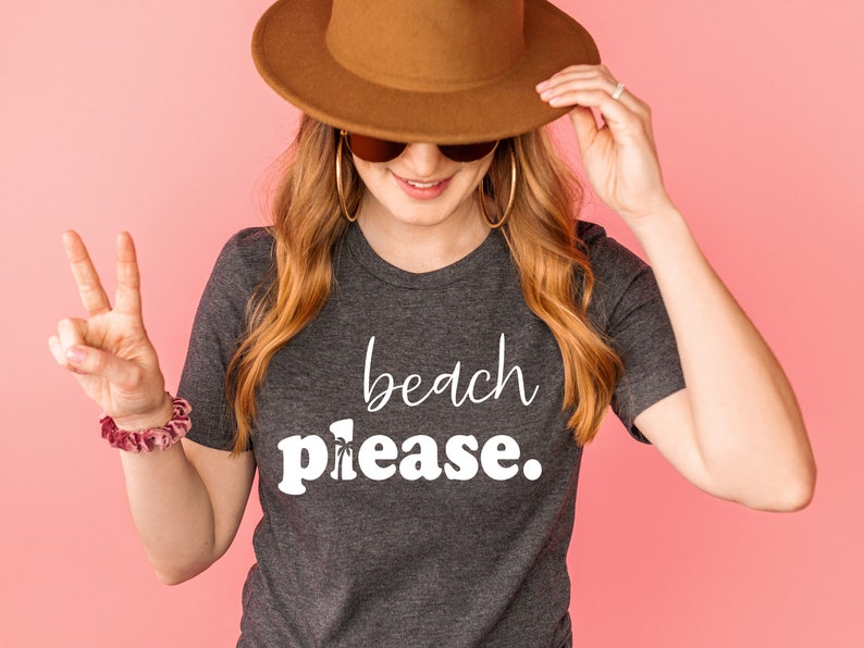 May include: A dark grey t-shirt with the words "beach please" in white script. The person in the image is wearing a brown hat, sunglasses, and gold hoop earrings. A scrunchie is on the wrist.