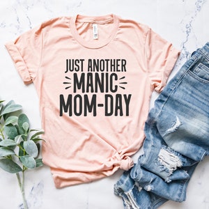 May include: A light pink t-shirt with the text "Just Another Manic Mom-Day" printed in black.