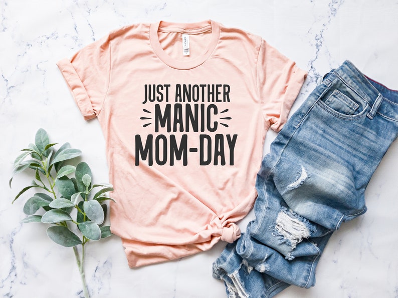 May include: A light pink t-shirt with the text "Just Another Manic Mom-Day" printed in black.