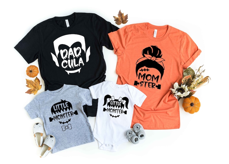 May include: A set of four Halloween-themed t-shirts for a family. The shirts are black, orange, gray, and white. The shirts feature the text "Dadcula", "Momster", and "Little Monster" with spooky graphics.