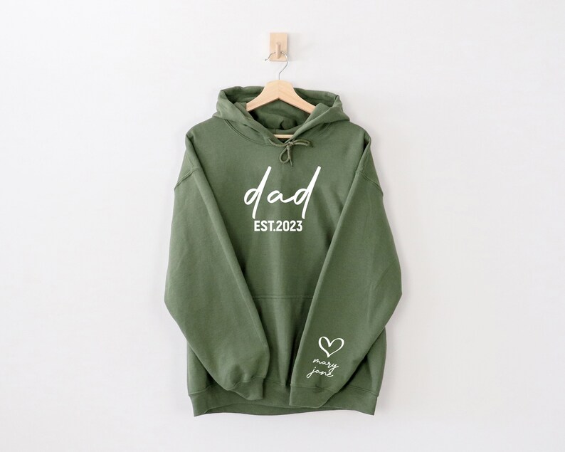 K&ouml;nnte beinhalten: Ein gr&uuml;ner Kapuzenpullover mit dem Text "dad est. 2023" und einem kleinen Herz mit dem Text "mary jane" auf dem &Auml;rmel.