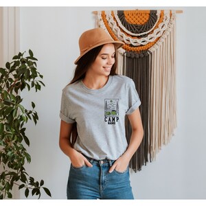 May include: A grey t-shirt featuring a camping scene graphic and the text "CAMP NAME". The shirt is being worn by a person with brown hair and a brown hat.