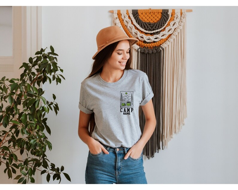 May include: A grey t-shirt featuring a camping scene graphic and the text "CAMP NAME". The shirt is being worn by a person with brown hair and a brown hat.