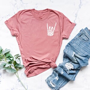 May include: A dusty rose t-shirt with a white skeleton hand making the rock and roll sign. The t-shirt is laying on a white marble surface with a pair of light blue distressed jeans.
