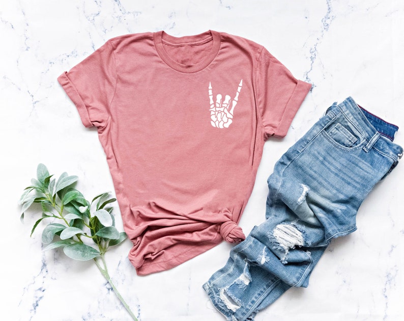 May include: A dusty rose t-shirt with a white skeleton hand making the rock and roll sign. The t-shirt is laying on a white marble surface with a pair of light blue distressed jeans.