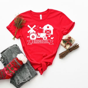 May include: A red t-shirt with a white graphic featuring a tractor, windmill, and barn, with the text "FARMGIRL" on a banner. The shirt is styled with folded sleeves, jeans, and a patterned sock.