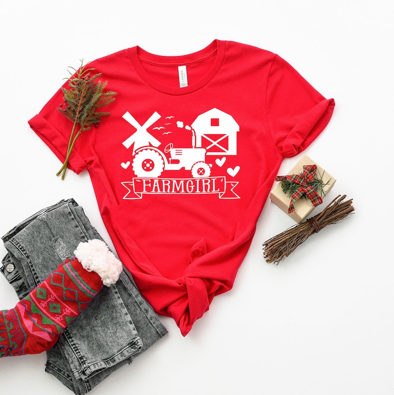 May include: A red t-shirt with a white graphic featuring a tractor, windmill, and barn, with the text "FARMGIRL" on a banner. The shirt is styled with folded sleeves, jeans, and a patterned sock.