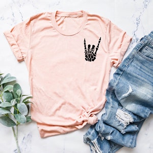 May include: A light pink t-shirt with a black skeleton hand making the rock and roll sign. The shirt is laid out on a white marble surface with a pair of blue jeans and a sprig of green foliage.