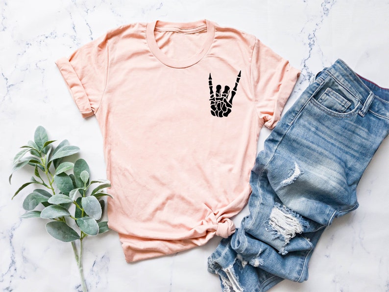 May include: A light pink t-shirt with a black skeleton hand making the rock and roll sign. The shirt is laid out on a white marble surface with a pair of blue jeans and a sprig of green foliage.