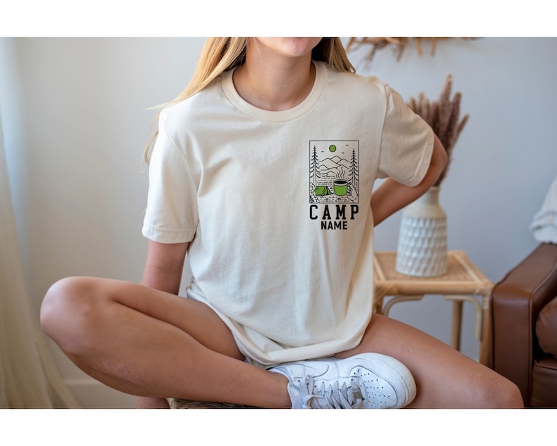 May include: A cream coloured t-shirt with a graphic of a camping scene with a coffee mug and a tent. The text "CAMP NAME" is below the graphic.