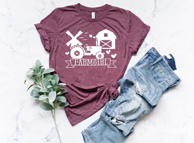 May include: A maroon t-shirt with a white graphic of a tractor, windmill, and barn, with the text "FARMGIRL" on a banner. The shirt is paired with blue jeans. A sprig of green leaves is in the lower left corner.