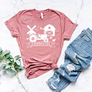 May include: A dusty rose t-shirt with a white graphic that says "FARMGIRL". The graphic includes a tractor, windmill, barn, and hearts. Light blue denim jeans with distressed details are folded next to the shirt.