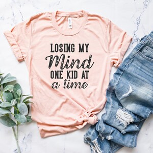 May include: A peach-colored t-shirt with the text "Losing My Mind One Kid At A Time" printed in black.