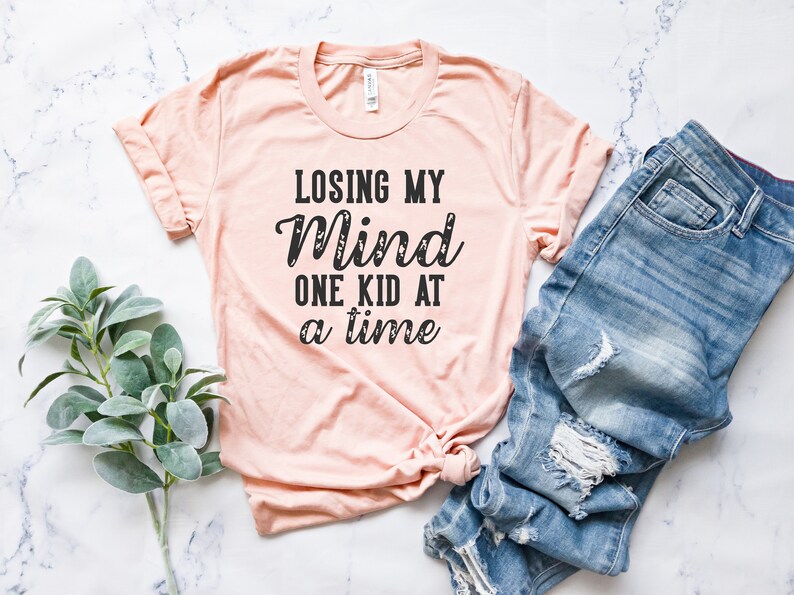 May include: A peach-colored t-shirt with the text "Losing My Mind One Kid At A Time" printed in black.