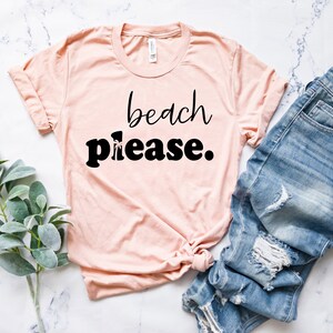 May include: A pale pink t-shirt with the words "beach please" in black script and bold font. The shirt is paired with blue denim jeans with distressed details. A sprig of green leaves is on the left.