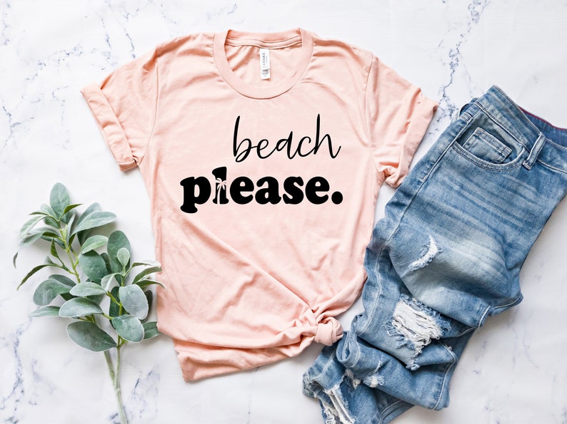 May include: A pale pink t-shirt with the words "beach please" in black script and bold font. The shirt is paired with blue denim jeans with distressed details. A sprig of green leaves is on the left.