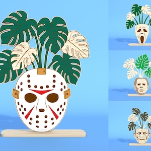 May include: A wooden decorative piece featuring a white Jason Voorhees mask with red accents and brown details, holding green and cream-colored monstera leaves. The mask is mounted on a light-colored wooden base, set against a blue background.