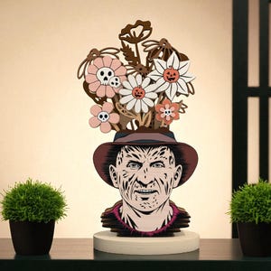 May include: A wooden sculpture of a character with a hat, adorned with flowers and skulls. The sculpture is painted in shades of brown, pink, and white, and is set on a white base. Two small potted plants flank the sculpture.