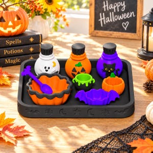 Peut inclure: Un plateau rectangulaire noir avec des motifs d'Halloween en relief contient trois petites bouteilles représentant un fantôme, une citrouille et un chat noir. L'ensemble inclut un bol orange en forme de citrouille, une cuillère violette et un chaudron vert.