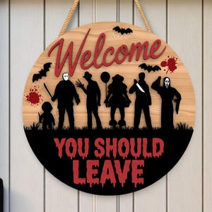 Puede incluir: Cartel redondo de Halloween de madera con las palabras "Welcome" y "YOU SHOULD LEAVE" en rojo. El cartel presenta siluetas de personajes de películas de terror, murciélagos y salpicaduras de sangre. Se adjunta una cuerda de yute para colgarlo.