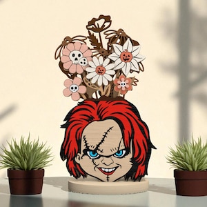 May include: A wooden decorative piece featuring a stylized Chucky face with red hair, blue eyes, and stitched scars. Above the face are wooden flowers in shades of pink, white, and brown. Two small potted plants flank the central figure.