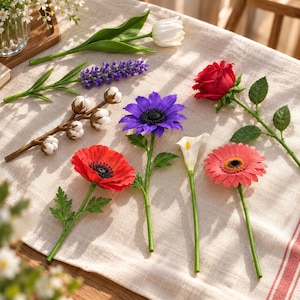 Puede incluir: Una colección de flores artificiales que incluye un tulipán blanco, lavanda morada, una rama de algodón, una rosa roja, una amapola roja, una margarita morada, un lirio de agua blanco y una gerbera rosa sobre una superficie de tela clara.