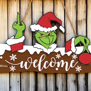 May include: Wooden welcome sign featuring a green character in a Santa hat, with a raised middle finger. The sign has a brown base with white snowflakes and the word "welcome" in white script. The character's arms and head are visible.