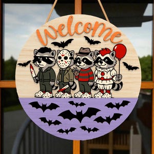 May include: A round wooden Halloween welcome sign featuring four cartoon raccoons dressed as horror movie characters. The sign has a light wood top with orange text and black bats. The bottom is purple with bat silhouettes.