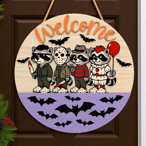 May include: Round wooden Halloween welcome sign featuring cartoon raccoons dressed as horror movie characters. The sign has a light wood grain background with orange "welcome" lettering and a purple lower section with bat silhouettes.