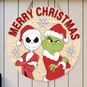 Może przedstawiać: Okrągły drewniany znak bożonarodzeniowy z Jackiem Skellingtonem i Grinchem w czapkach Świętego Mikołaja. Napis "MERRY CHRISTMAS" jest czerwony, z białymi płatkami śniegu. Znak jest zawieszony na beżowej linie.