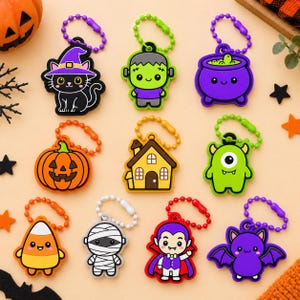 May include: A collection of nine Halloween-themed rubber keychains featuring a black cat in a purple hat, a green monster, a purple cauldron, an orange pumpkin, a brown house, a green cyclops, candy corn, a white mummy, a vampire, and a bat.