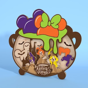 May include: Wooden Halloween-themed decoration in the shape of a cauldron. The cauldron features a green, orange, and purple bow, with a green slime effect. Inside are cartoon characters and the words "Hocus Pocus".