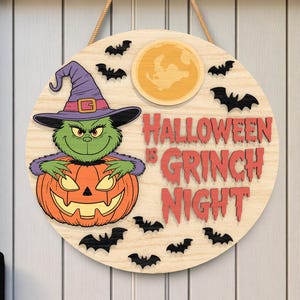 Puede incluir: Un letrero redondo de madera de Halloween con un personaje verde de Grinch con un sombrero de bruja y una calabaza naranja tallada. El letrero dice "Halloween is Grinch Night" en rojo, con murciélagos negros y una luna amarilla.