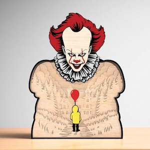 May include: A wooden layered art piece featuring a menacing clown with red hair and makeup, a white ruffled collar, and a sinister grin. Below, a child in a yellow raincoat holds a red balloon, set against a forest and house backdrop.