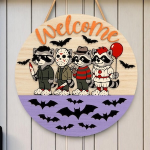May include: Round wooden Halloween welcome sign featuring cartoon raccoons dressed as horror movie characters. The sign has a light wood grain background with the word "Welcome" in orange. A purple section at the bottom has black bat cutouts.