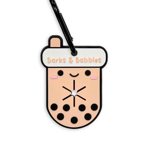 May include: A pink and white boba tea charm with a black clip. The charm has a cute cartoon face with black eyes and a pink blush. The text "Barks & Babbles" is written on the top of the charm.