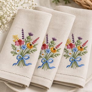 May include: Three folded beige linen napkins featuring an embroidered bouquet of colorful wildflowers including red, yellow, blue, and purple blossoms with green stems and leaves, tied together with a bright blue ribbon bow at the base.