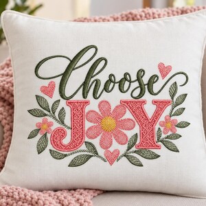 May include: A square off-white throw pillow featuring embroidered text that reads Choose JOY in dark green and coral pink thread. The design includes a central coral pink flower with a yellow center, surrounded by green leaves and small coral hearts.