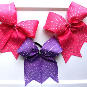 May include: Three cheerleading bows in vibrant colors. Two are hot pink, and one is purple. Each bow is adorned with sparkling silver rhinestones arranged in a dotted pattern. The bows are made of a shiny satin material.