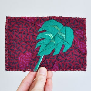 Puede incluir: Una hoja de tela verde sobre un fondo burdeos. La hoja está hecha de muchas pequeñas piezas de tela cosidas juntas. El fondo es una tela burdeos con un patrón de pequeñas formas negras y moradas.