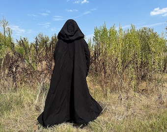 May include: A black hooded cloak with a flowing design. The cloak is worn outdoors in a field of tall grass and plants under a blue sky. The cloak has a hood and drapes down to the ground.