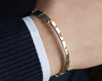 Op de afbeelding: Een gouden en zilveren kabelarmband met een rij kleine roze edelstenen. De armband zit om de pols van een persoon.