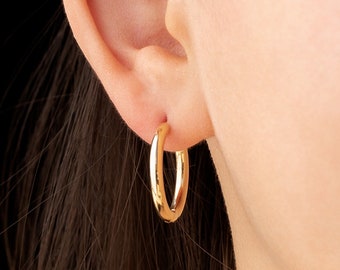May include: A close-up of a gold hoop earring worn on an ear. The earring is a simple, polished gold, with a rounded shape. The background is black, which highlights the earring's color and shape.
